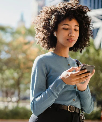 Woman with earbuds walking while looking at her phone.
